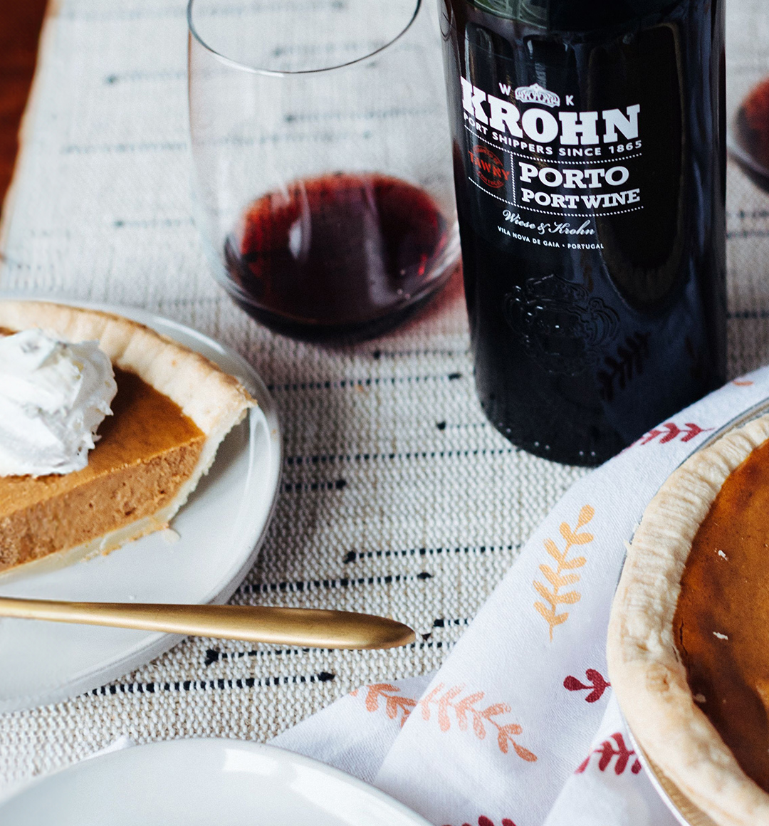 A bottle of port wine on a table next to a stemless wine glass filled with port and a plate with a slice of pumpkin pie.