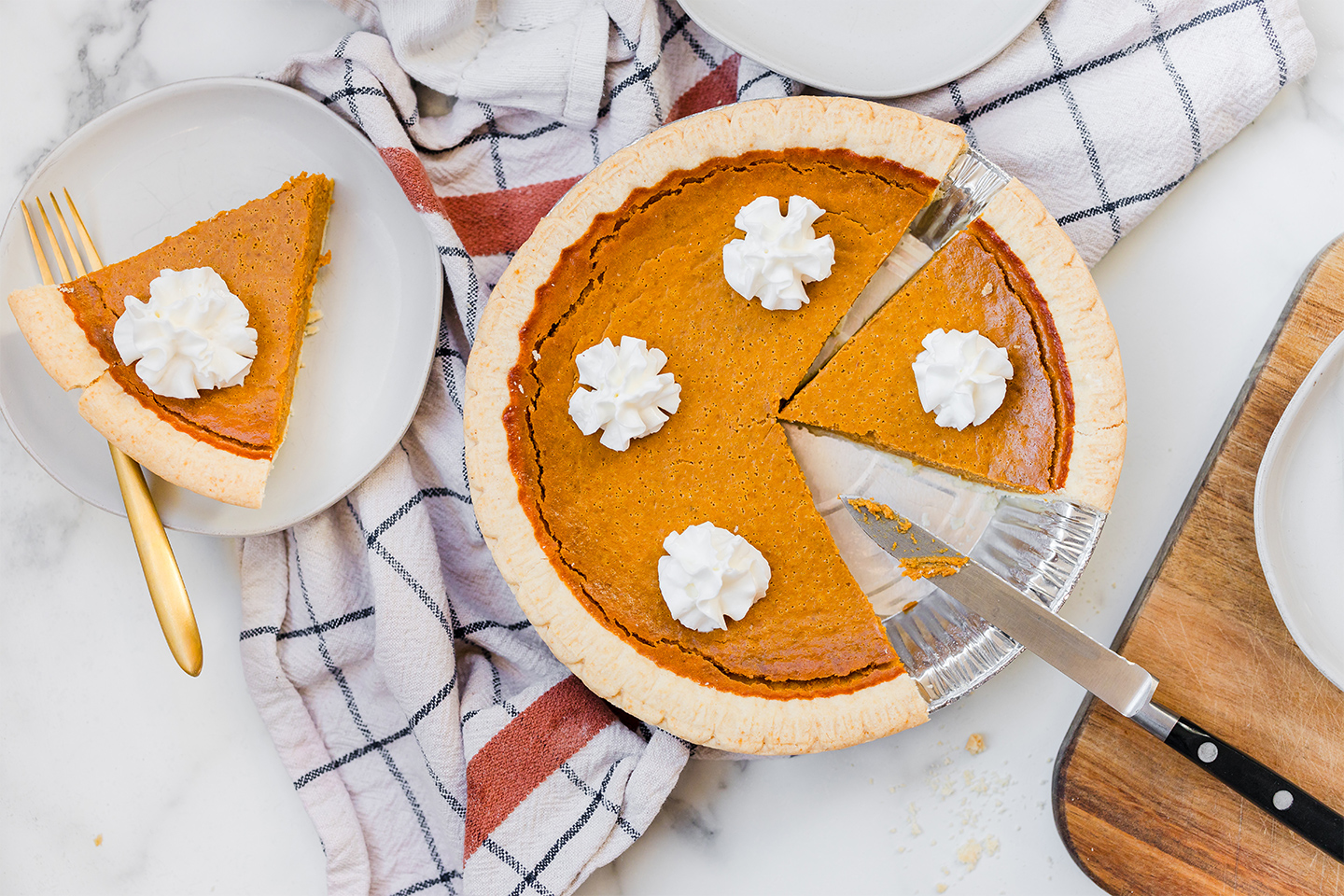 An overview image of a pumpkin pie and whipped cream with a slice taken out and placed on a plate beside it. 