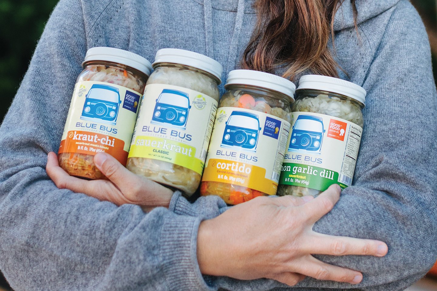 A person in a grey sweatshirt holding 4 jars of Blue Bus saurkraut.