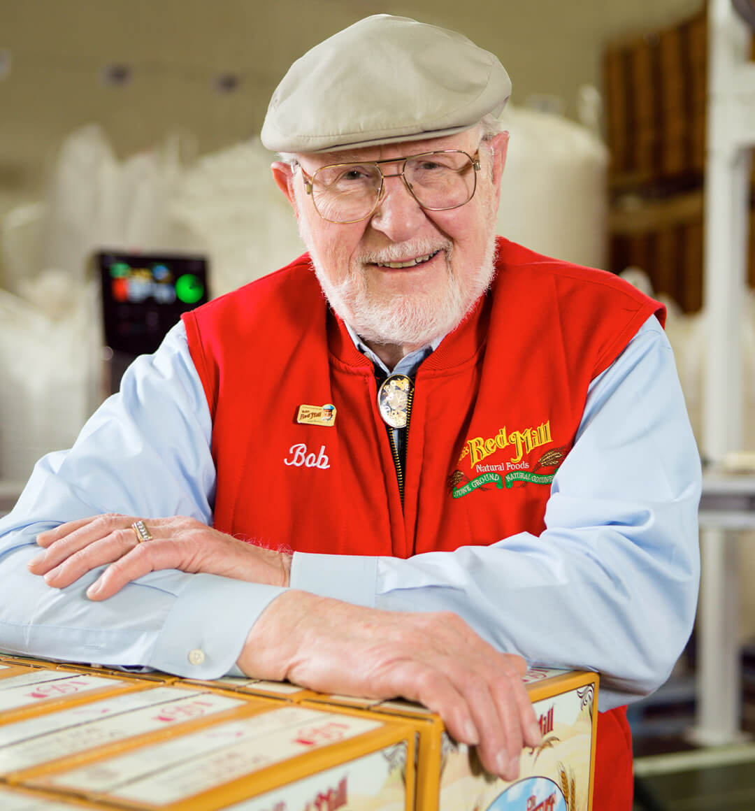 a portrait of Bob, the founder of Bob's Red Mill. He is wearing glasses, a blue longsleeve shirt and red vest.