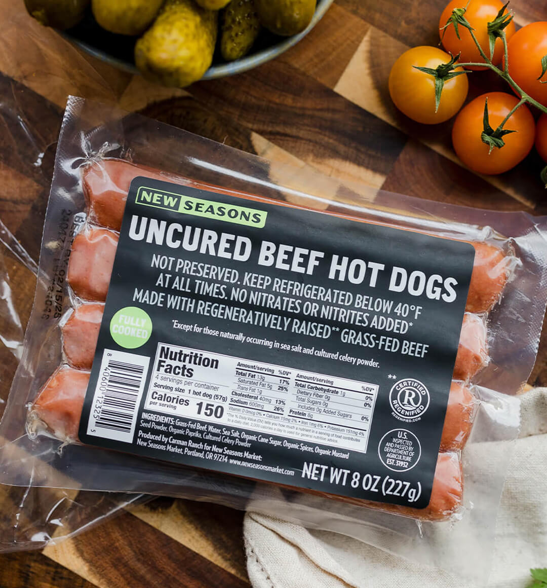 a package of New Seasons uncured beef hot dogs on a wood board with tomatoes in the background.