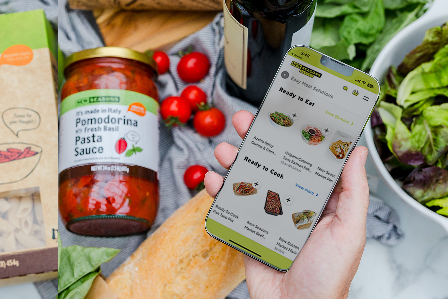 A hand holding a phone featuring an online shopping app with a tabletop in the background displaying a jar of marinara sauce, a box of dried pasta, a glass of wine, and a salad in a bowl.