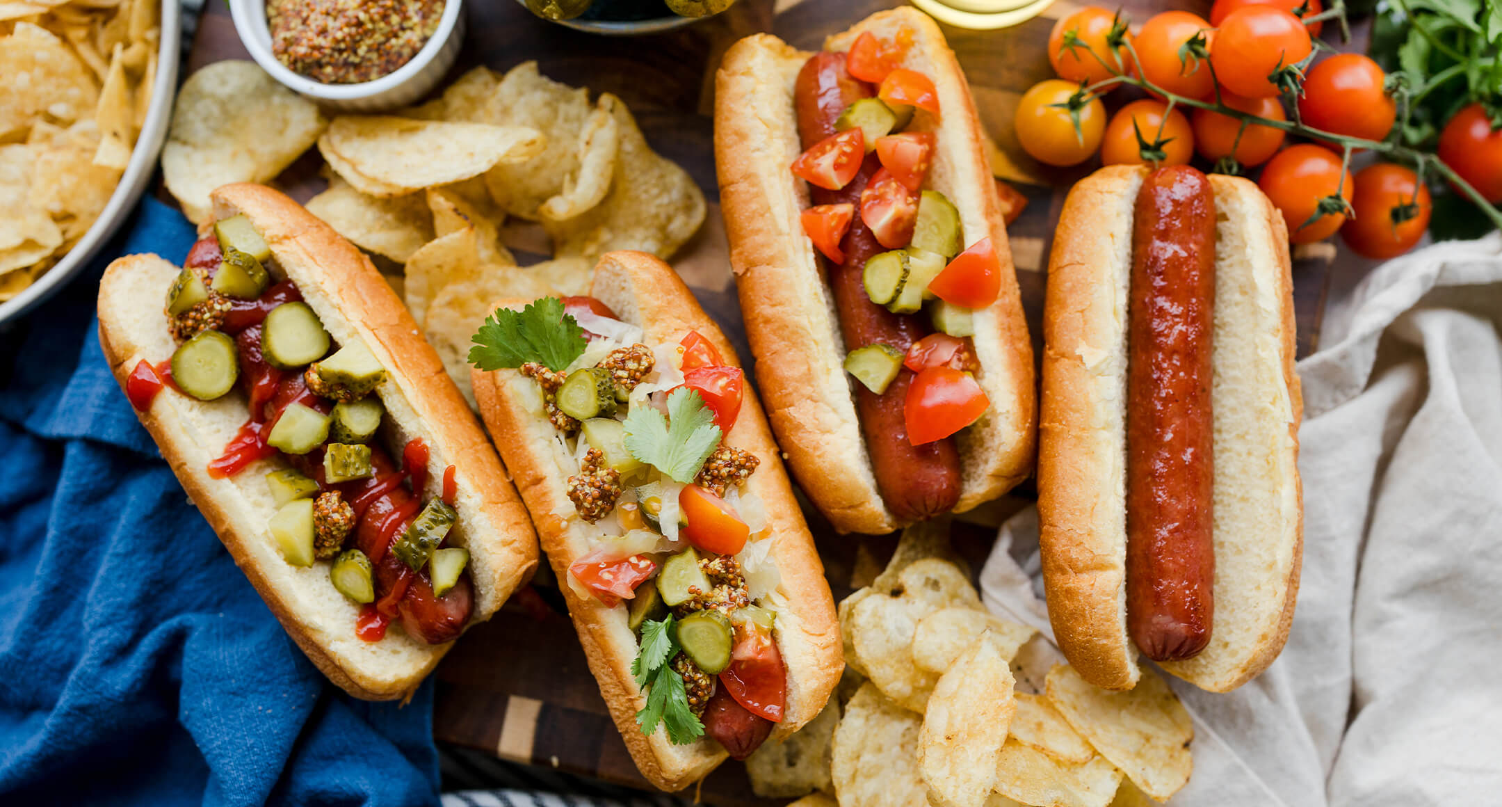 Four New Seasons Uncured Beef Hot Dogs in buns with mustard, chopped tomatoes, and pickles and chips on the side. 