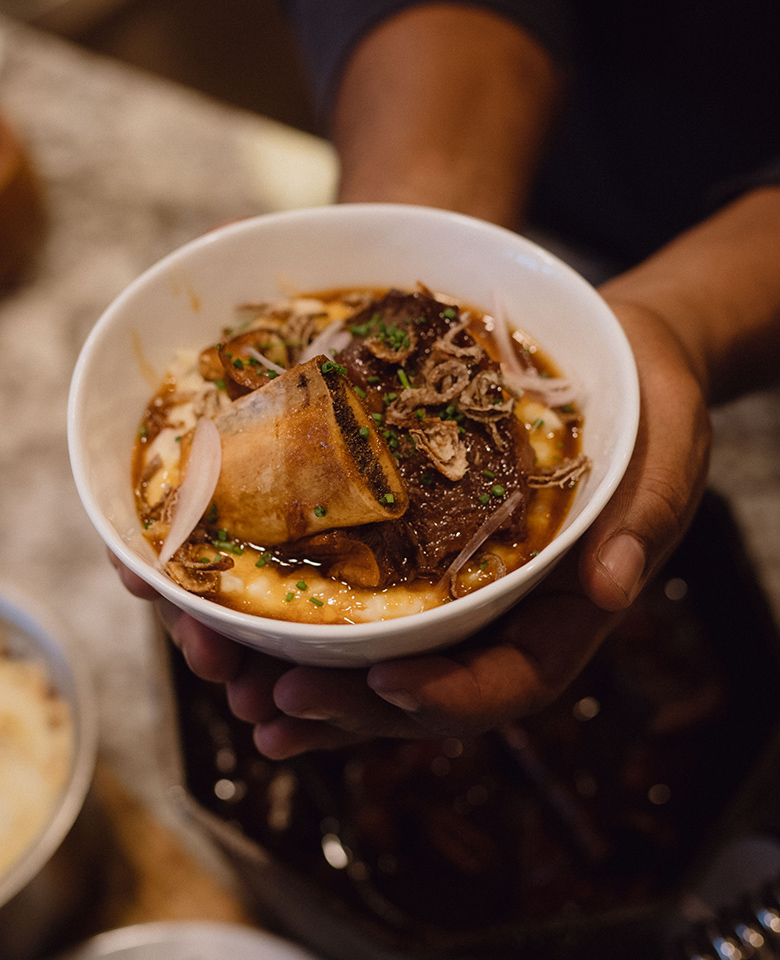 A close-up of two hands holding a bowl filled with mashed potatoes topped with slow-braised short ribs. 