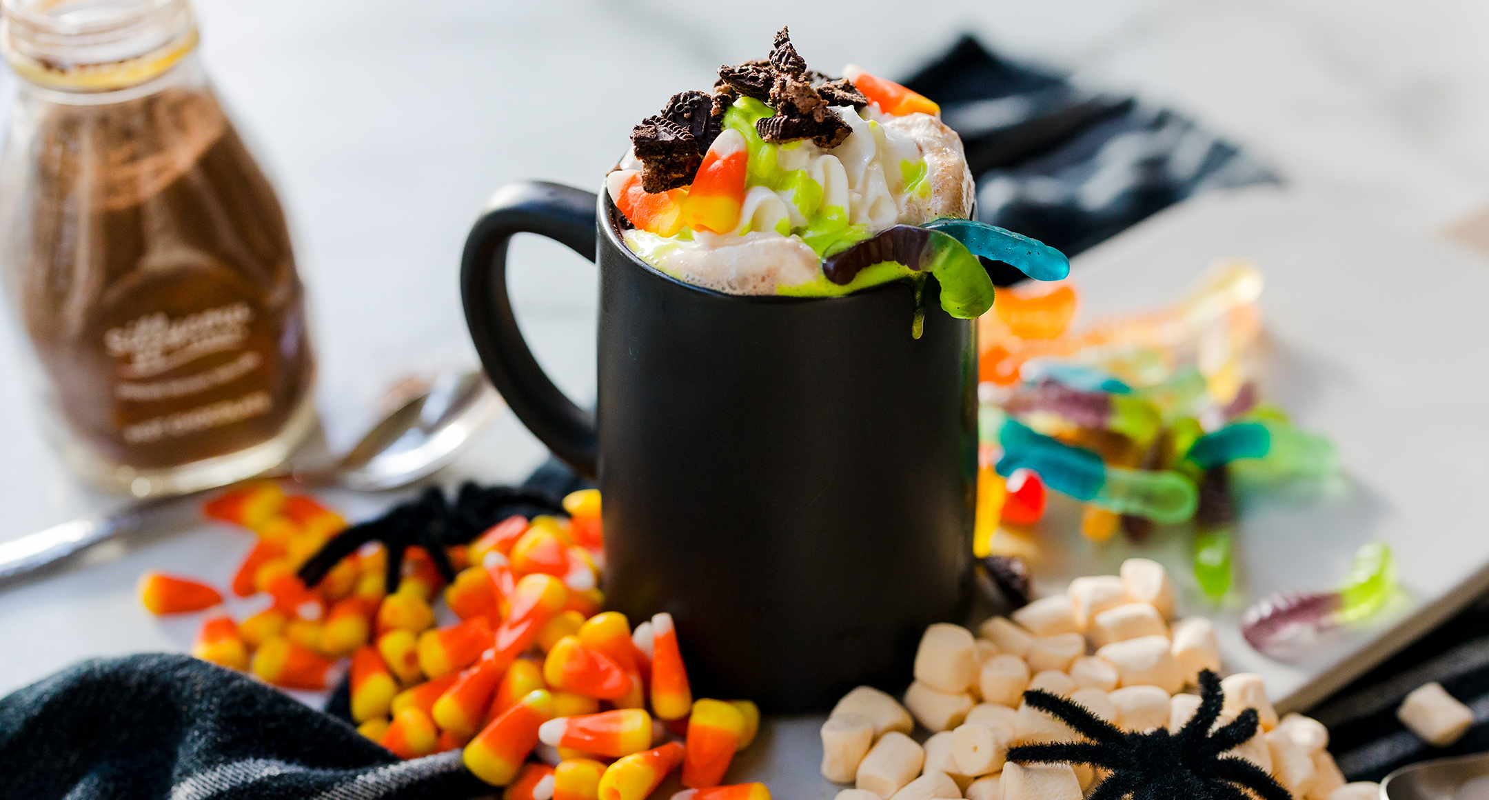 A black mug filled with hot chocolate and topped with festive green whipped cream, gummy worms, candy corn, and marshmallows.