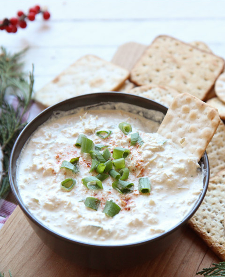A bowl of artichoke dip surrounded by a variety of crackers. 