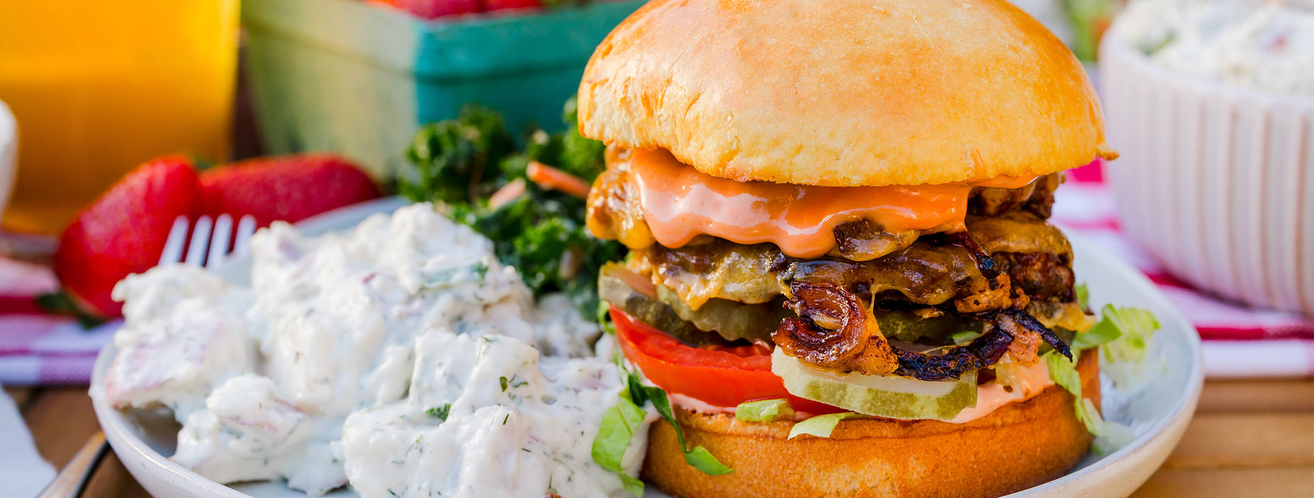 a burger on a bun piled high with cheese, lettuce, tomato and more in a table scene with potato salad and more..