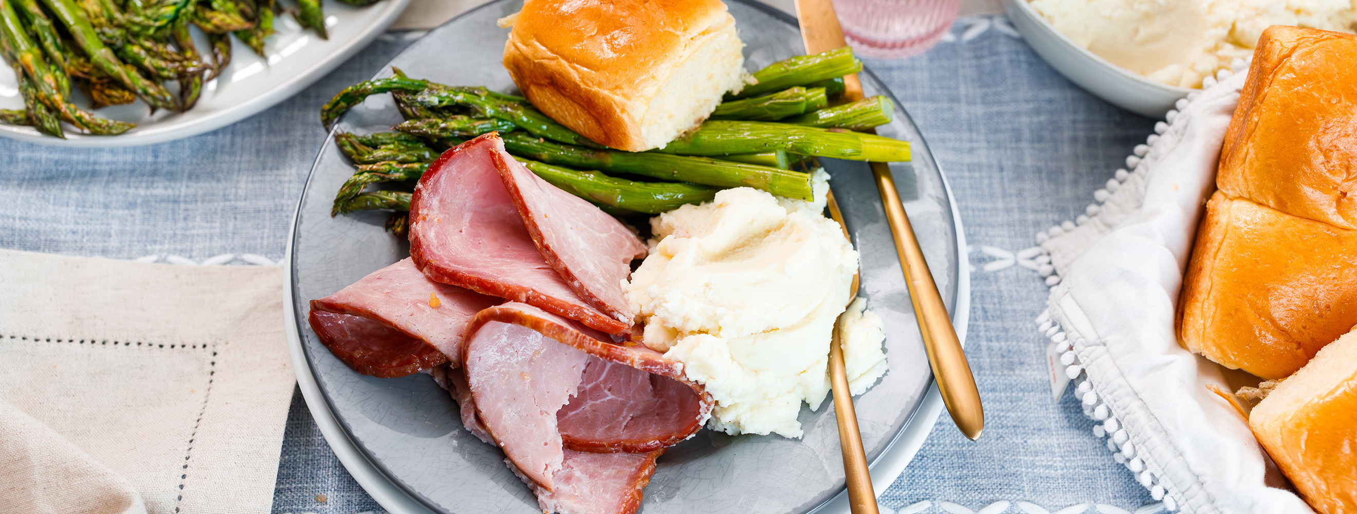 a plated Easter dinner with sliced ham, mashed potatoes, asparagus and rolls.
