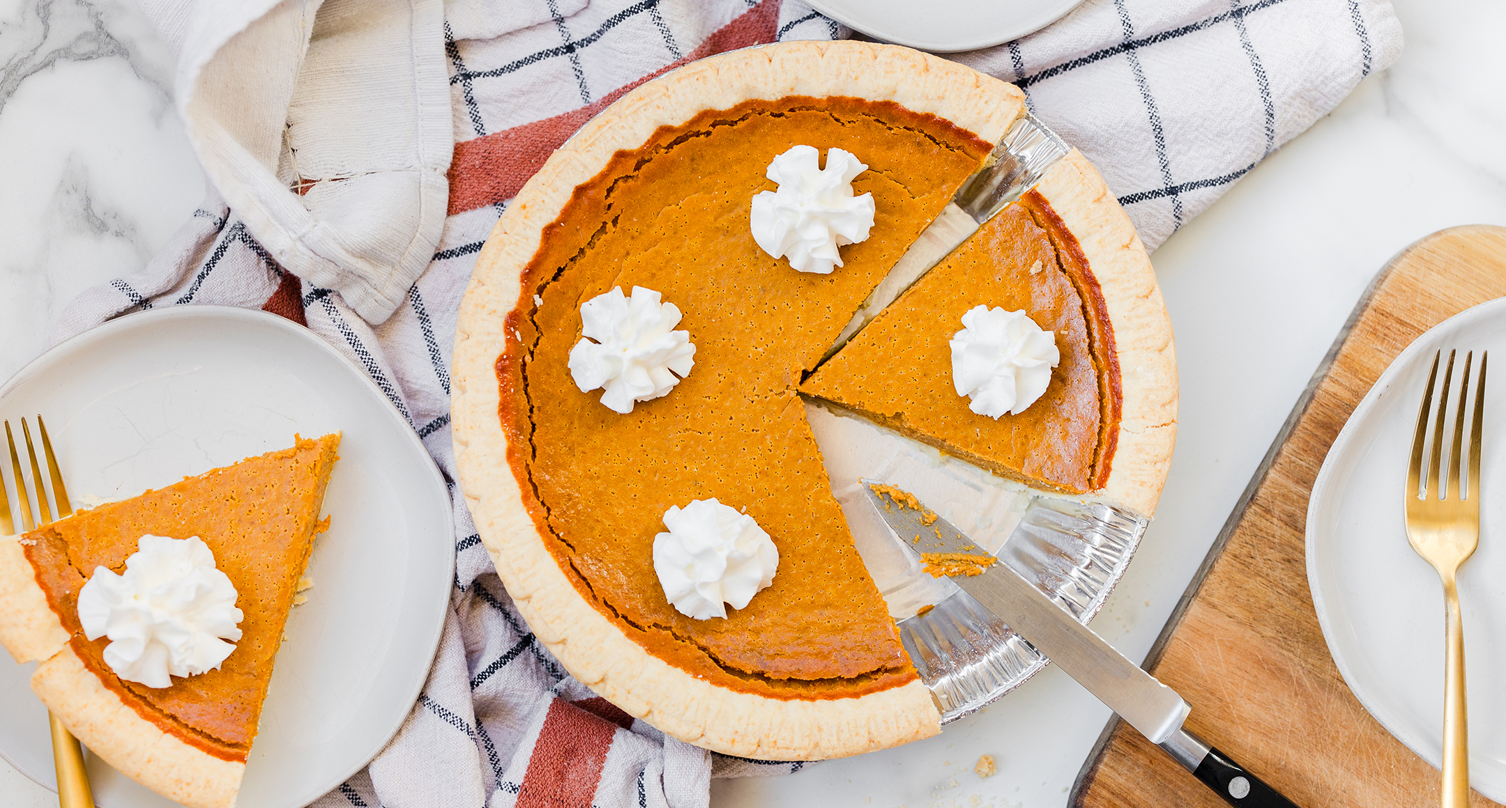 A freshly baked pumpkin pie topped with whipped cream and sliced with one piece removed and placed on a plate next to it.