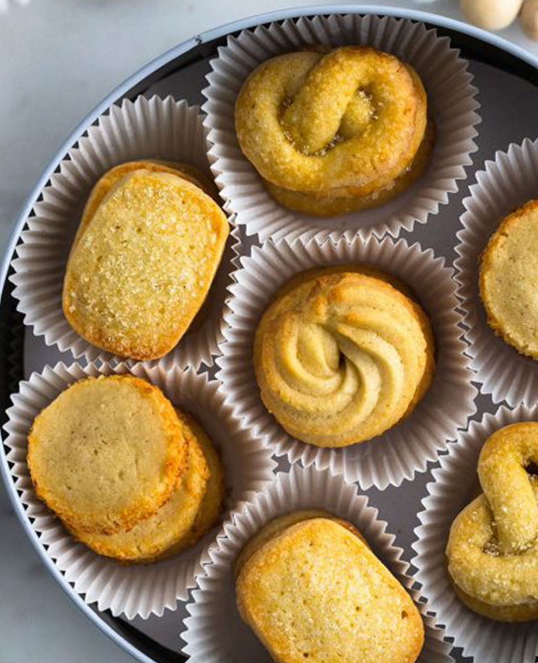 Butter cookies in four different shapes placed in paper cupcake liners inside a cookie tin. 