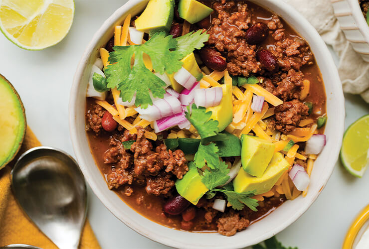 A bowl of beef chili topped with sliced avocado and shredded cheese.