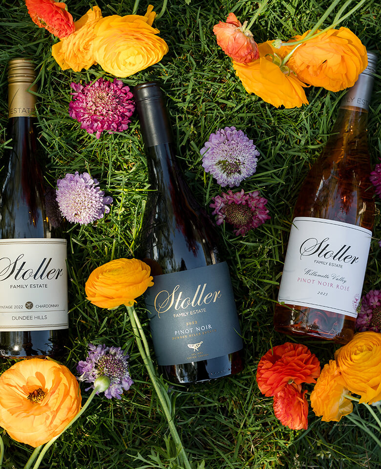 bottles of Stoller Family Estate wine lying in grass and colorful flowers.