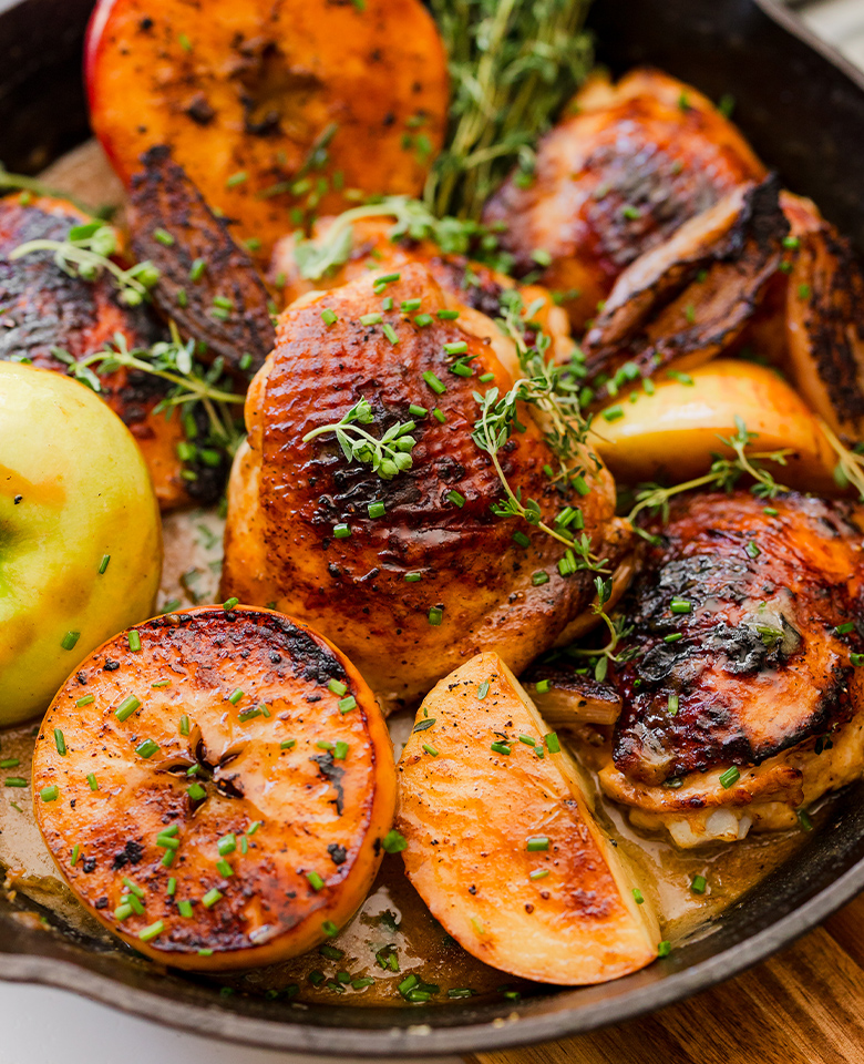 A cast-iron pan with braised chicken thighs, halved apples, shallots and herbs.