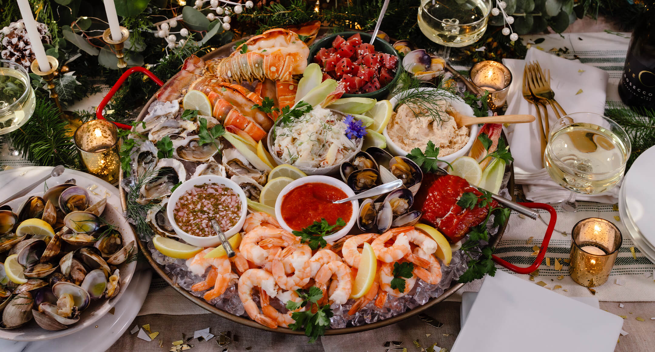 A holiday seafood spread, including cooked shrimp and lobster tails, raw clams, seafood dip, and more served on ice.