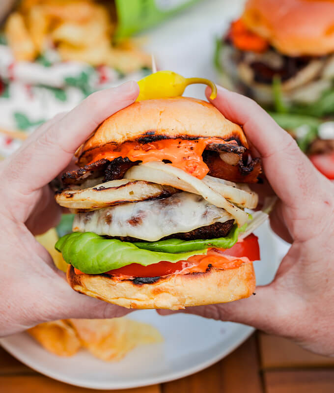 Two hands holding a burger with cheese, tomatoes, grilled onions, lettuce and Calabrian Chili mayo