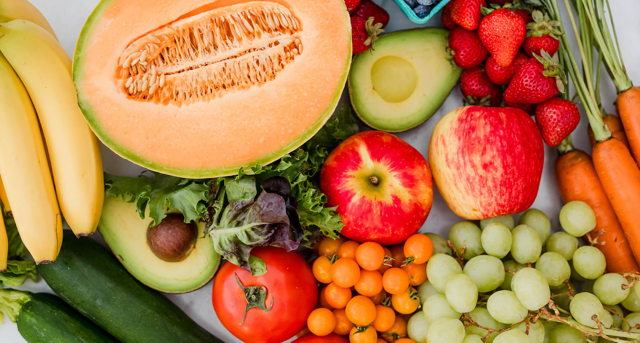 An array of fresh fruits and veggies, including half of a cantaloupe, carrots, and strawberries.