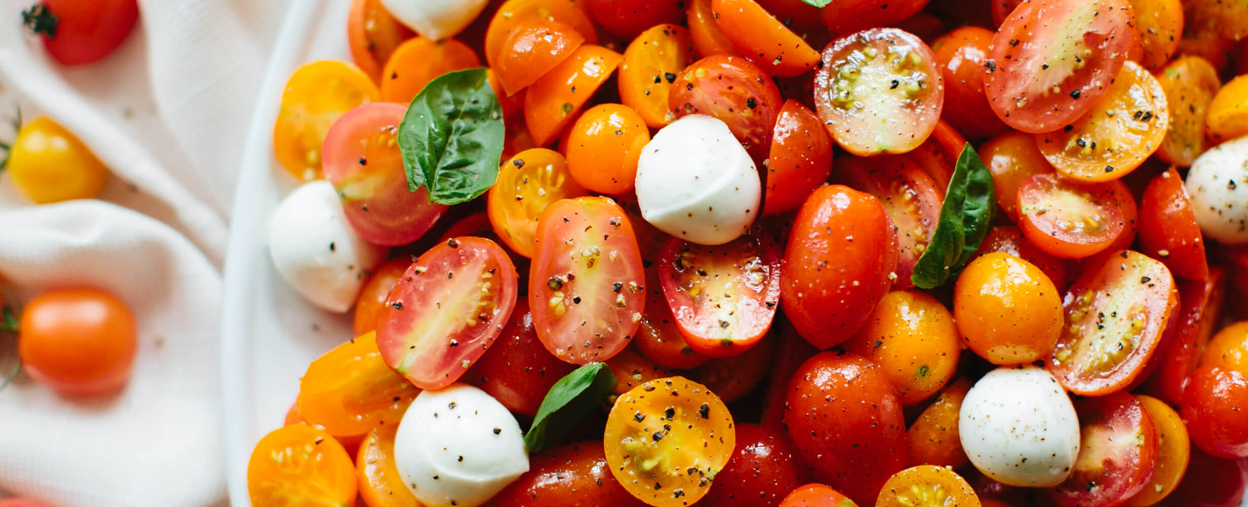 An image of cut up local tomatoes on a plate with fresh mozzarella and basil. Sprinkled with pepper.