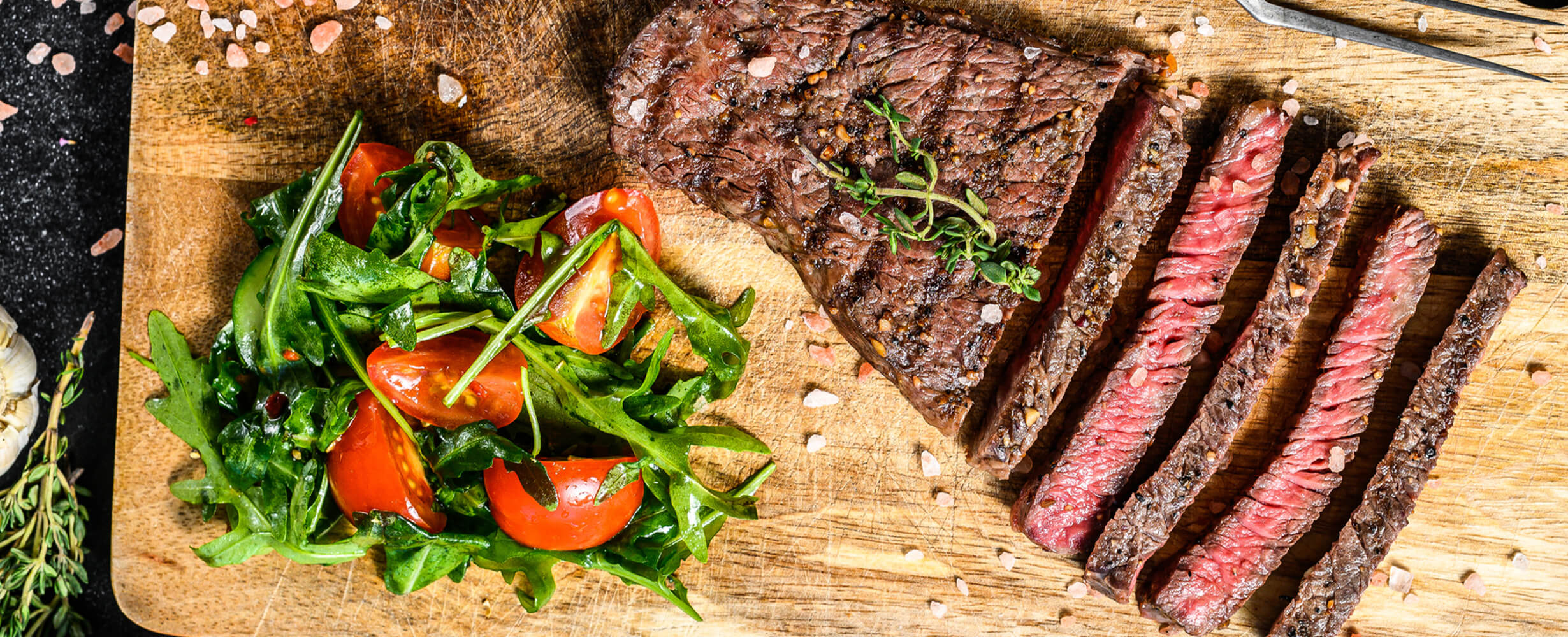 An image of a sliced sirloin steak on a wood board with tomatoes and greens.