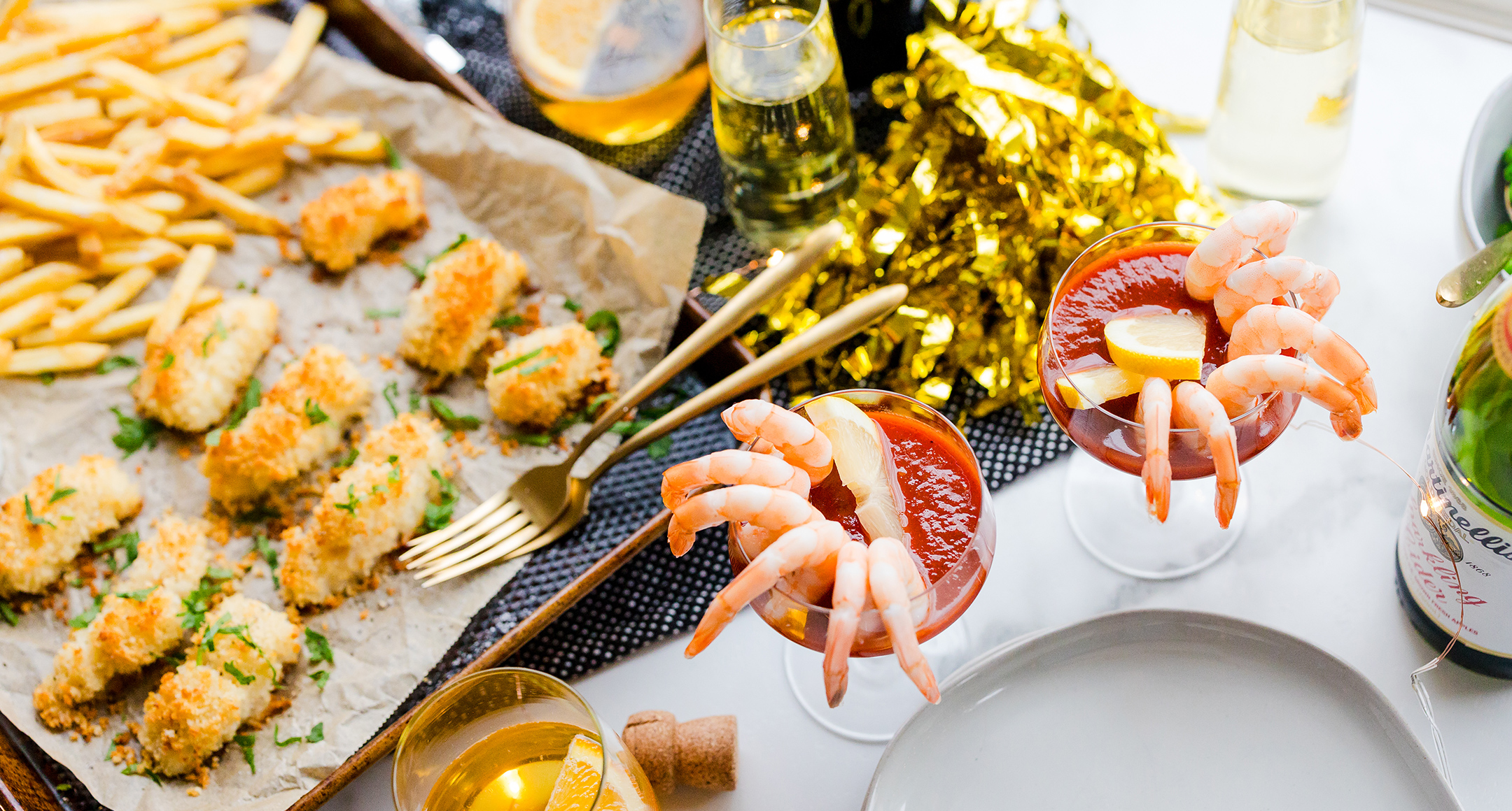 A party spread featuring sheet-pan fish & chips, shrimp lining a glass filled with cocktail sauce, and a glass of champagne.