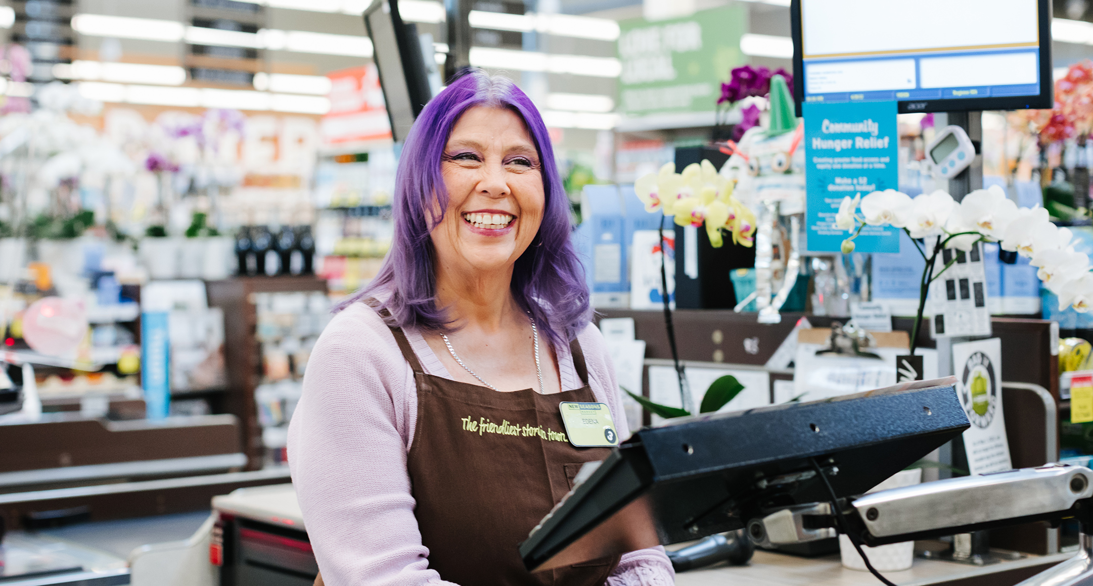 Smiling cashier at New Seasons Market checkout with grocery store view in the background. 