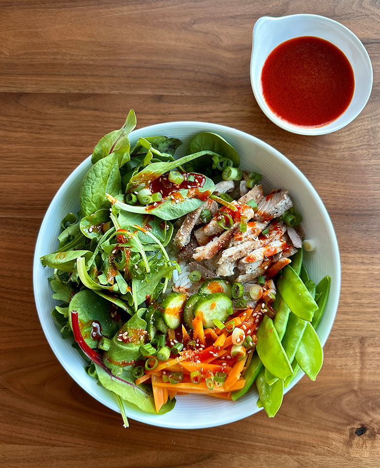 A rice bowl with fresh vegetables and pork, and a bowl of gochujang sauce on the side.