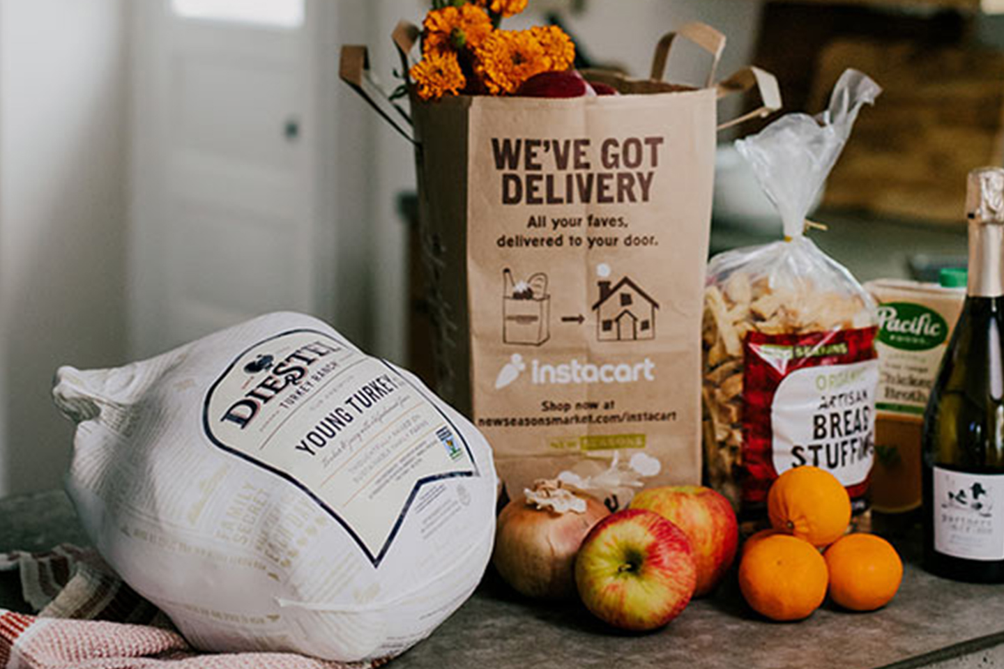 A New Seasons Market grocery bag on a table with bagged bread stuffing and a turkey with wine and fruit.