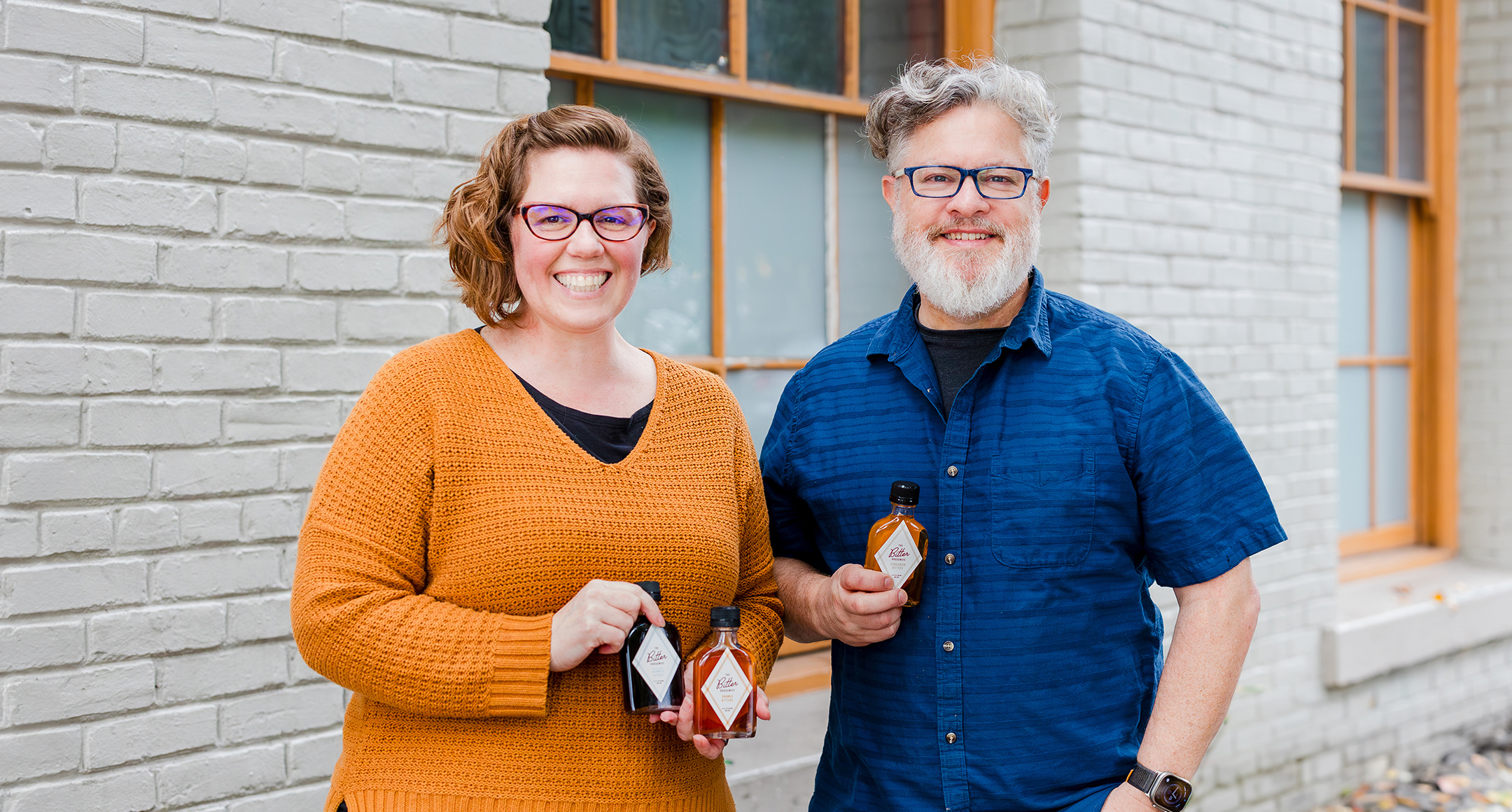 The photo features Genevieve and Dan Brazelton, owners of The Bitter Housewife, holding their Orange Bitters. 