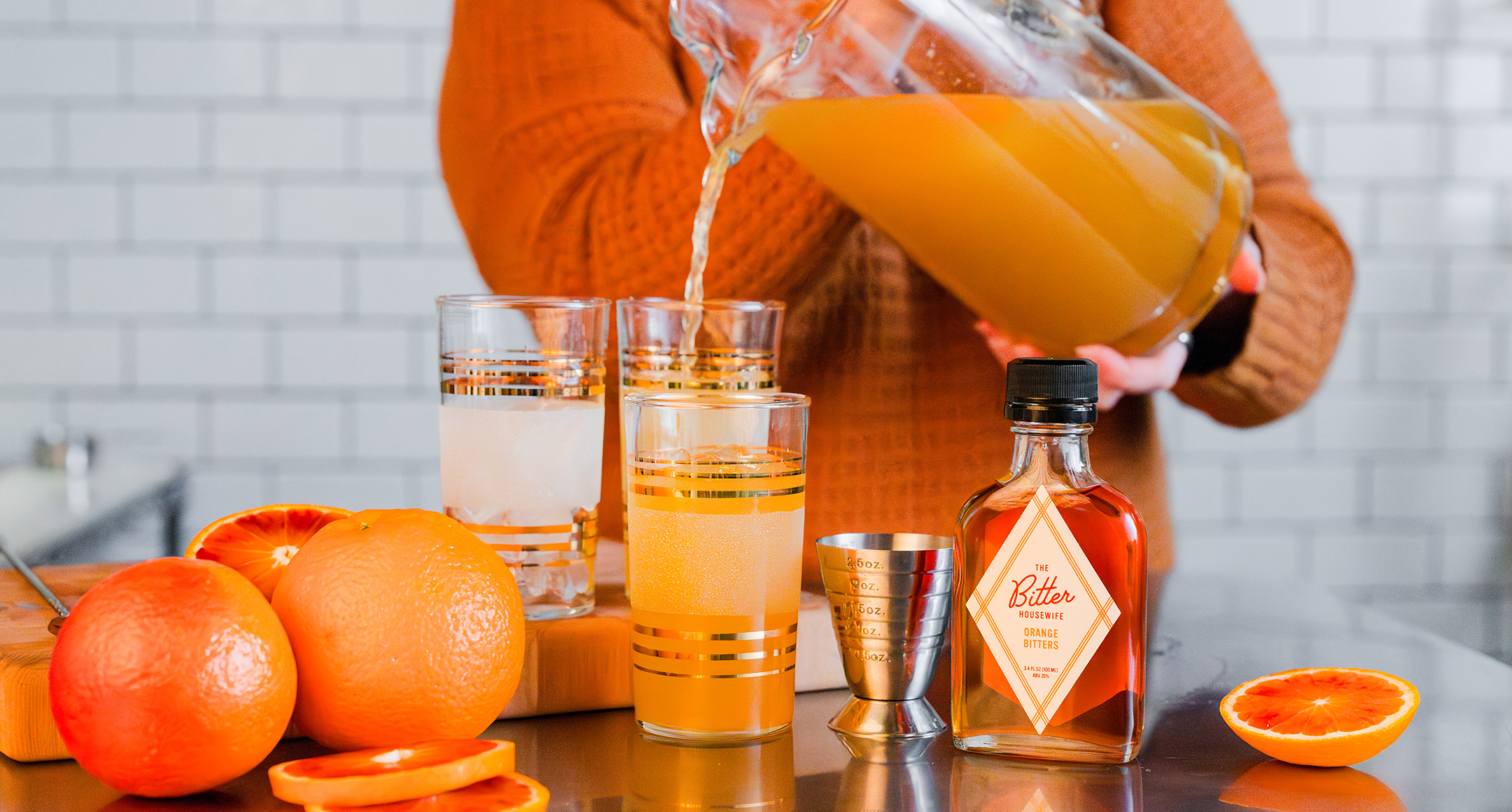 A table set with The Bitter Housewife Orange Bitters, two glasses being filled from a pitcher, and fresh oranges on the side. 