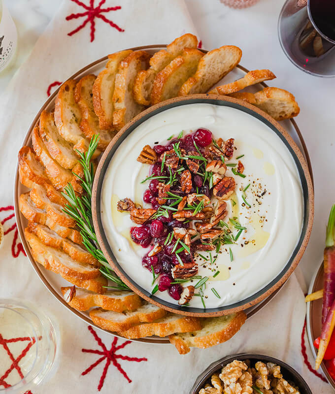 a beautiful picture of a holiday dip topped with cranberry and nuts and surrounded by sliced baguette