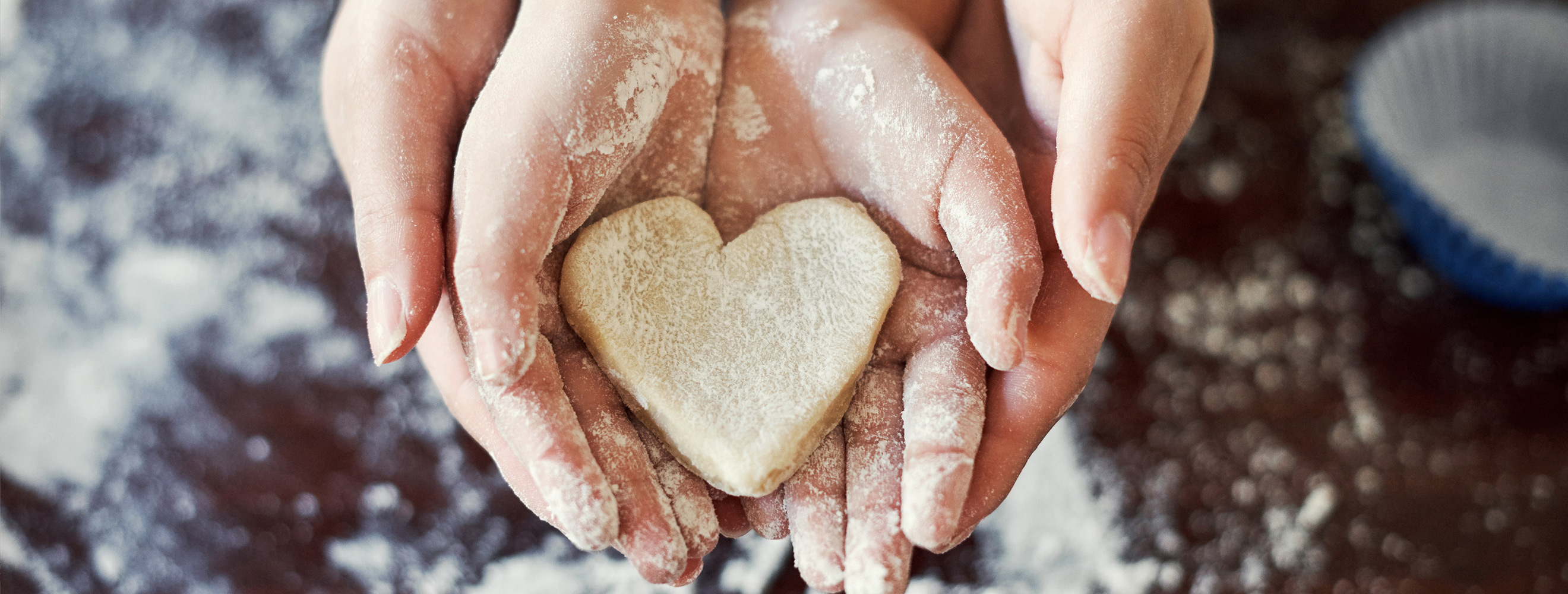 two sets of hands holding a heart-shaped piece of cookie dough above a floured surface