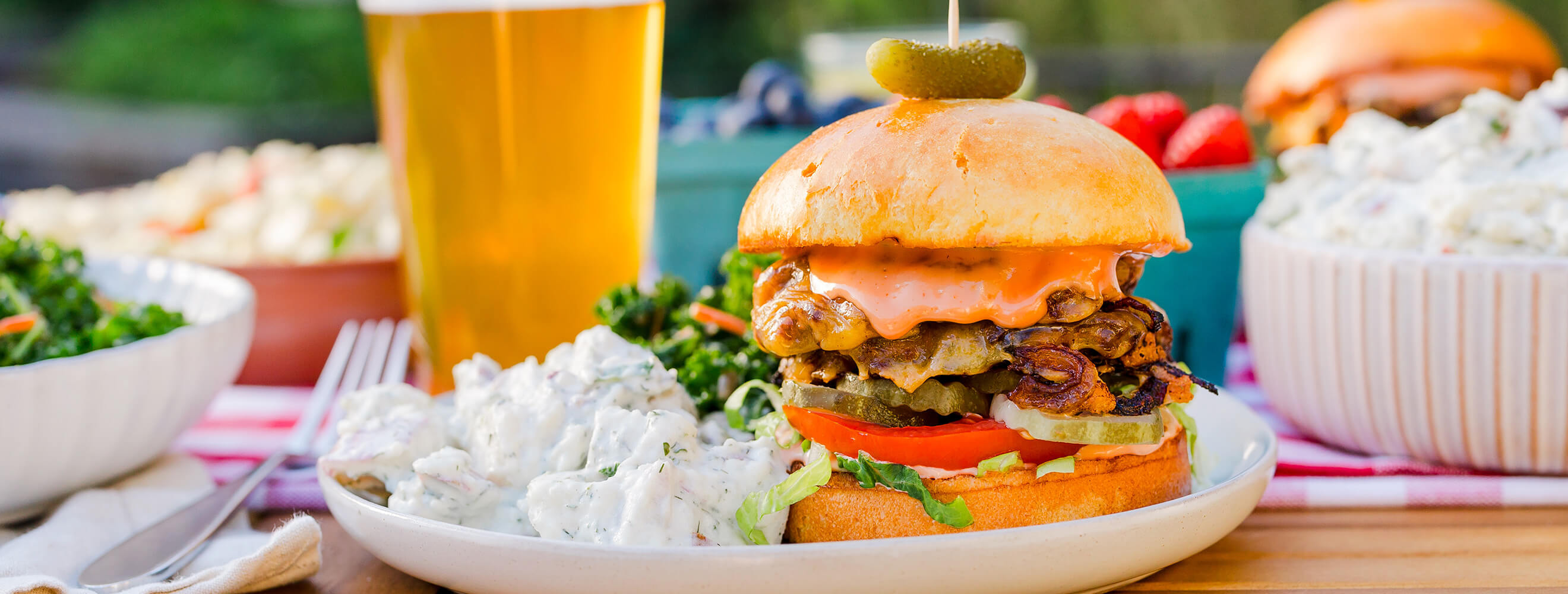 a hamburger on a plate with a side of potato salad and a glass of beer in the background.