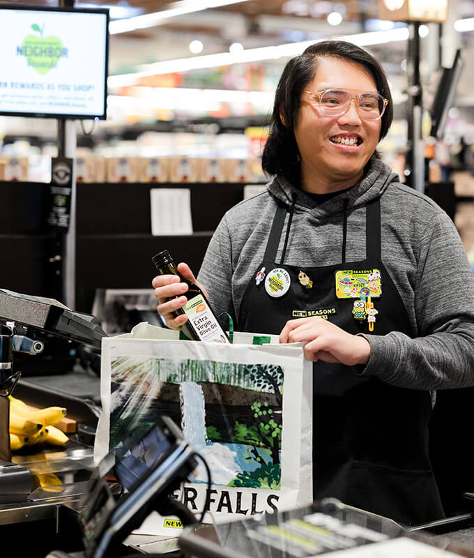a smiling person bagging groceries in a reusable bag at a grocery cash register.