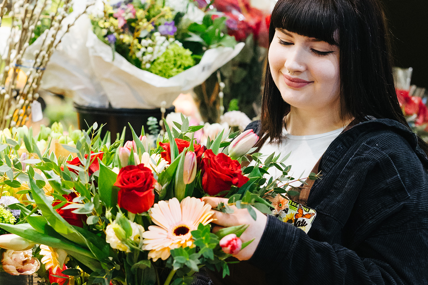 A New Seasons florist is smiling while they arrange a bouquet of roses and daisies.