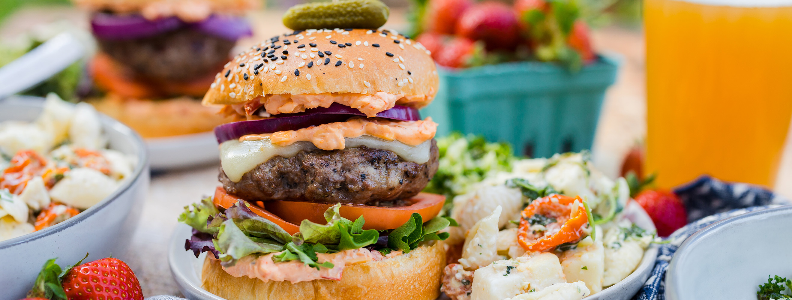 a burger on a bun piled high with cheese, lettuce, tomato and more in a table scene with deli sides and fresh fruit.