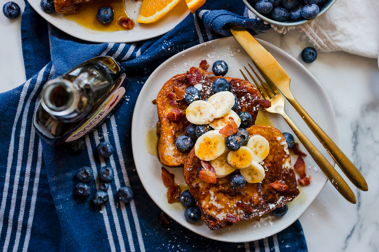 brioche french toast covered in fresh fruit and maple syrup on a plate with fork and knife.