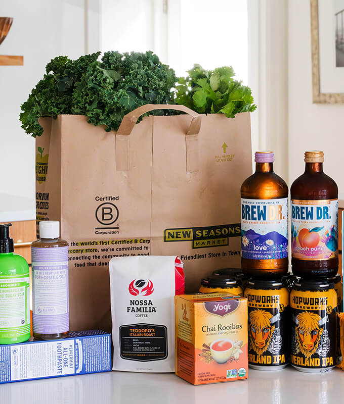 Straight on shot of an assortment of items including Yogi Chai Rooibos Tea, Nossa Familia Coffee, Brew Dr Kombucha, Hopworks IPA, and a brown New Seasons bag with the B Corp logo.
