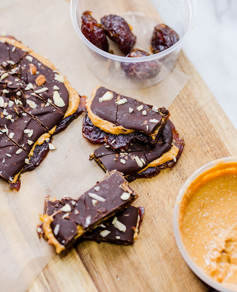 Date snickers bars on a wooden surface next to a container of dates and another container of nut butter.