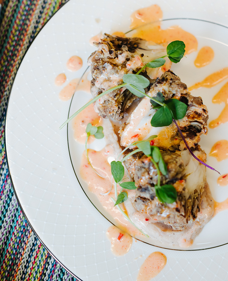 A close-up image of a plate with Maitake schnitzel served atop a white bean garlic hummus and drizzled with harissa aioli. 