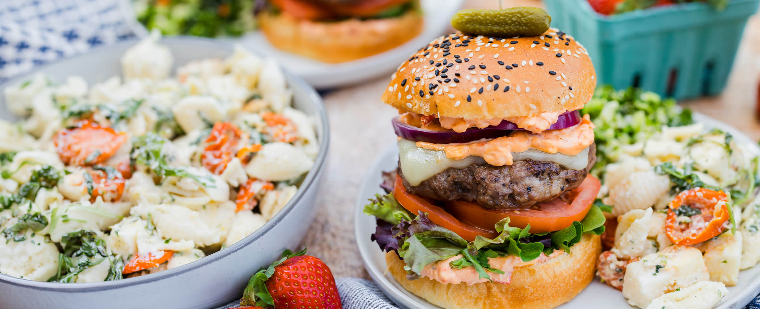 A ground beef burger with all the fixings on a plate with pasta salad.