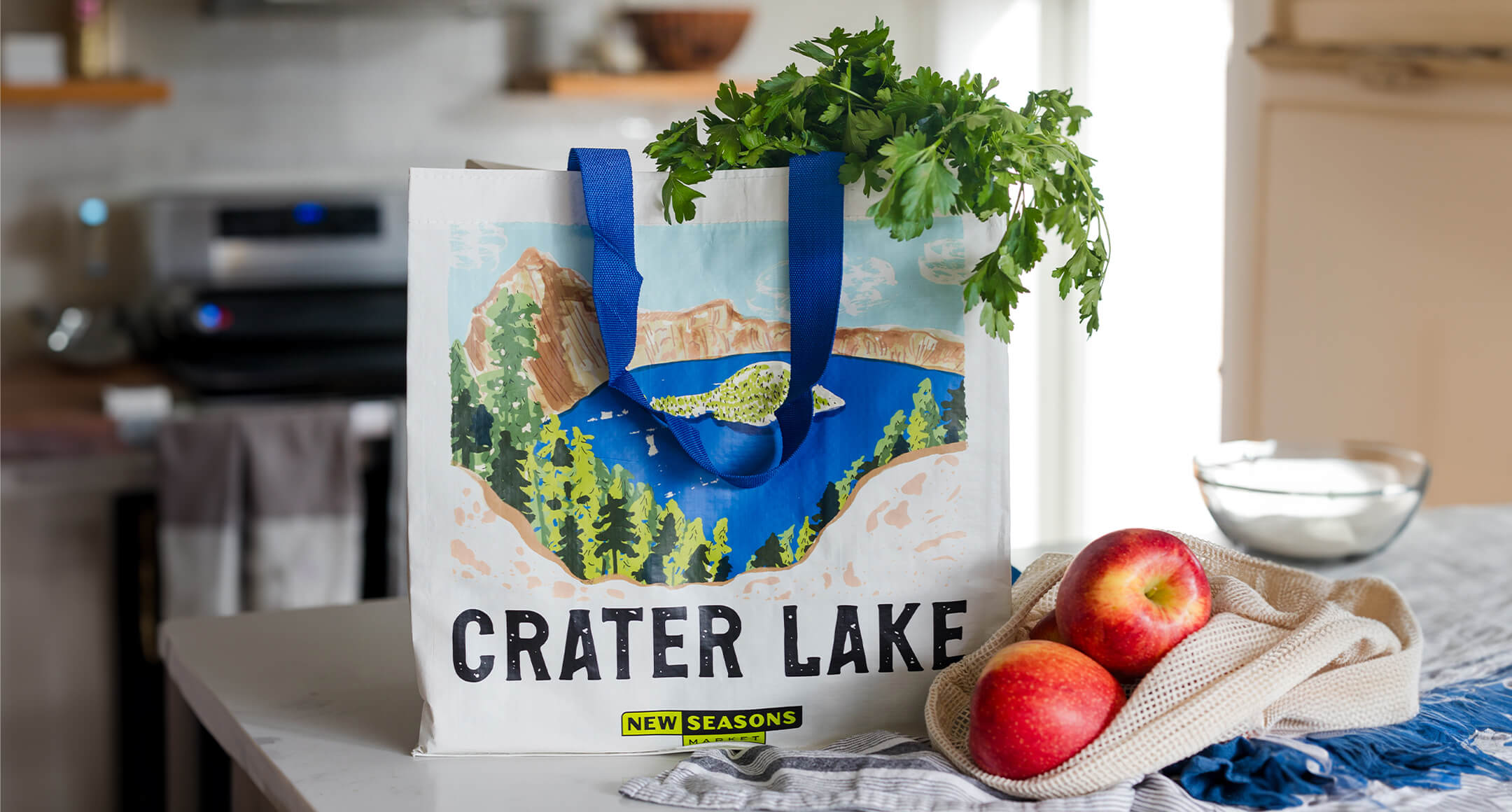 A reusable grocery bag sitting on a counter featuring an artistic depiction of Crater Lake National Park on the side.