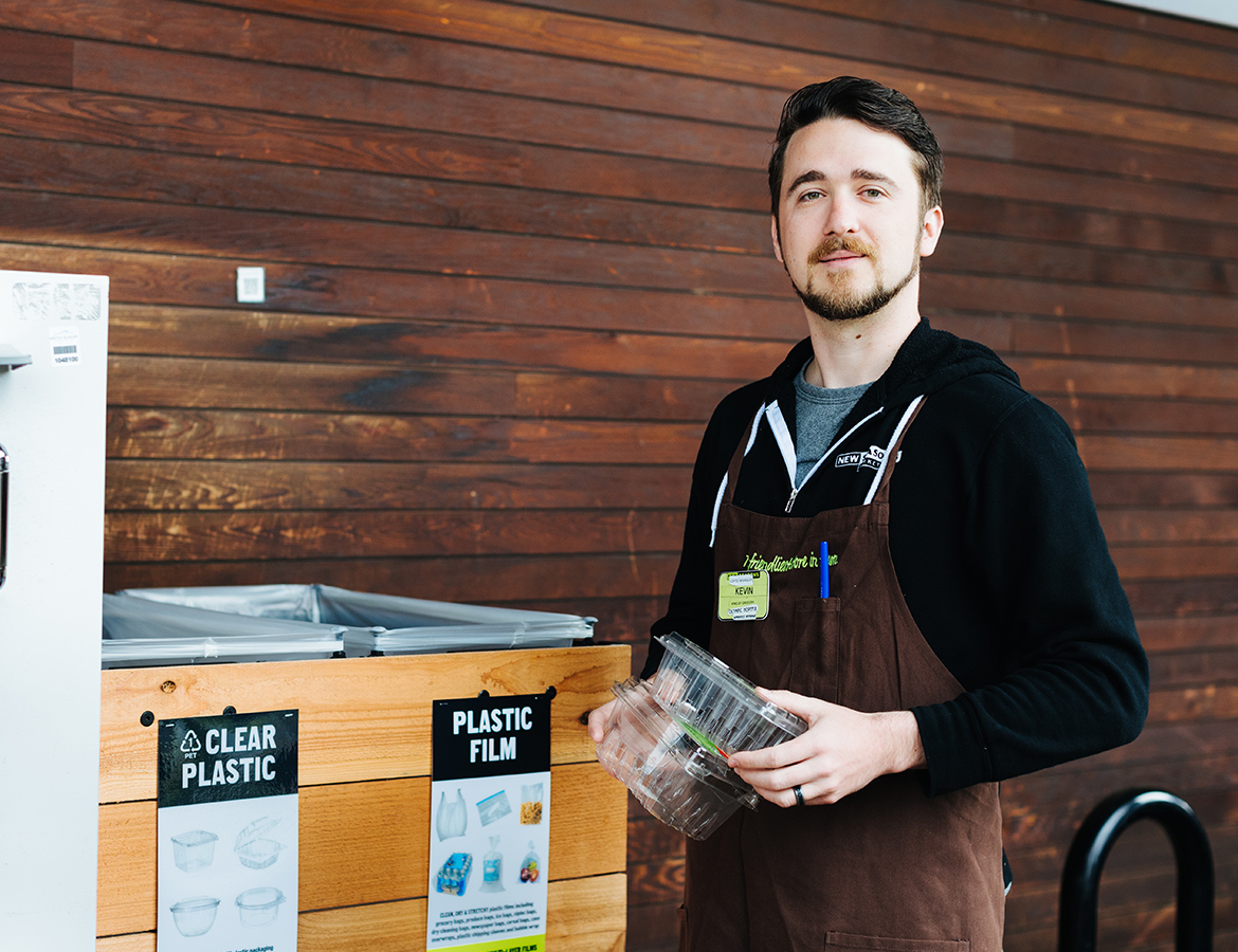 New Seasons Market staff member in a brown apron recycling plastic at a store.