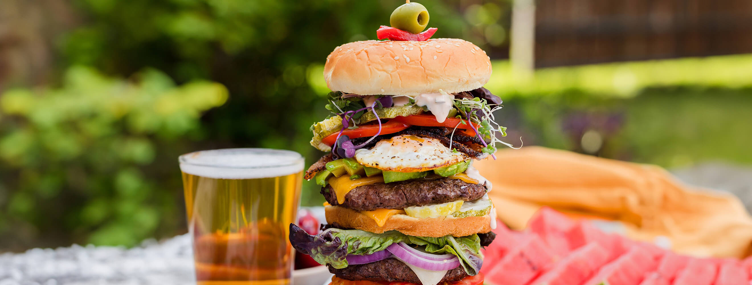 a burger stacked high with layers of meat, cheese, bread and vegetables in a picnic scene.
