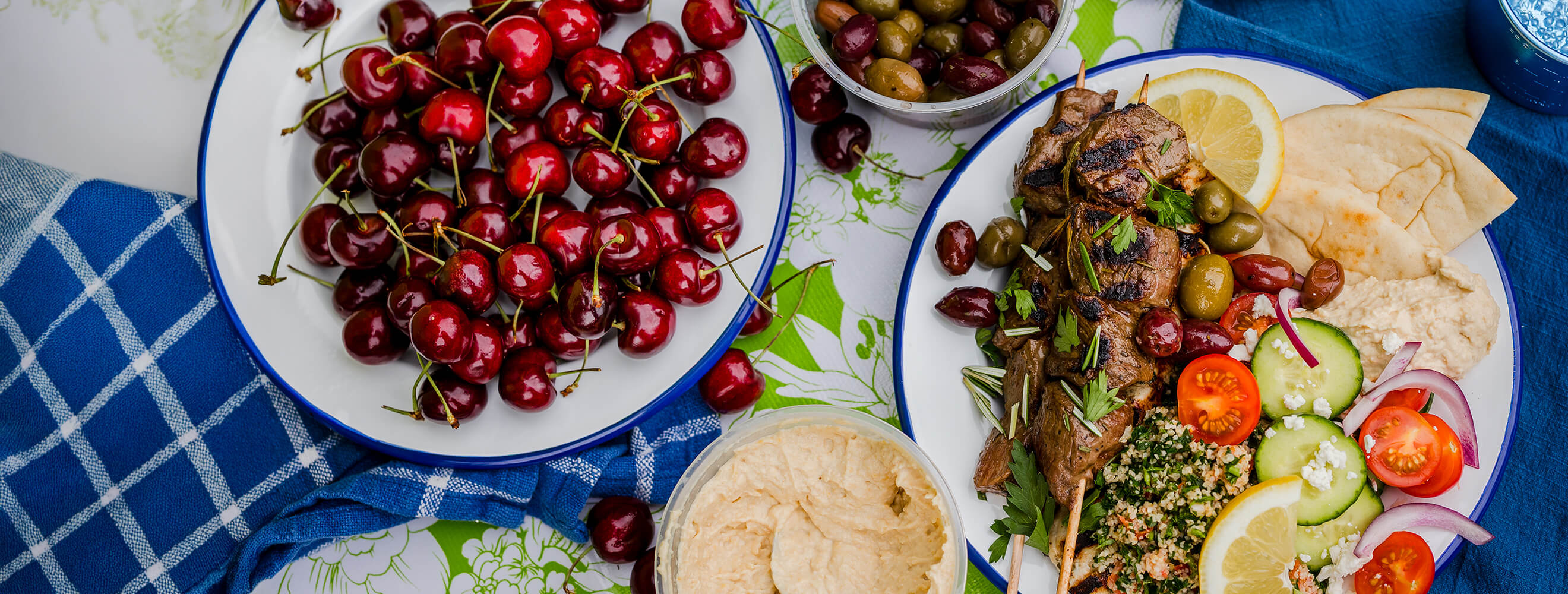Grilled beef skewers plated with pita, hummus, tabouleh, marinated olives, sliced cucumber and cherry tomatoes