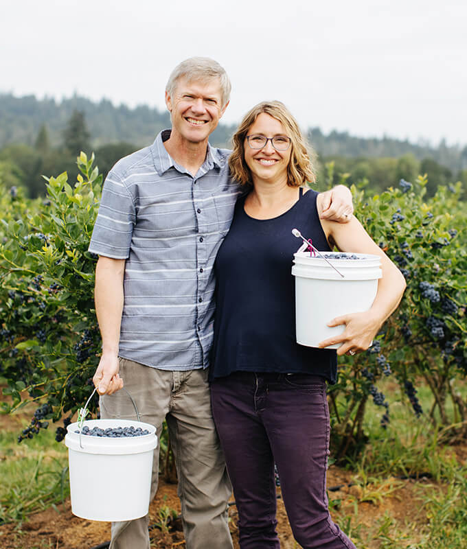 two smiling people standing in a field of blueberry bushes holding bucket full of fresh whole blueberries.
