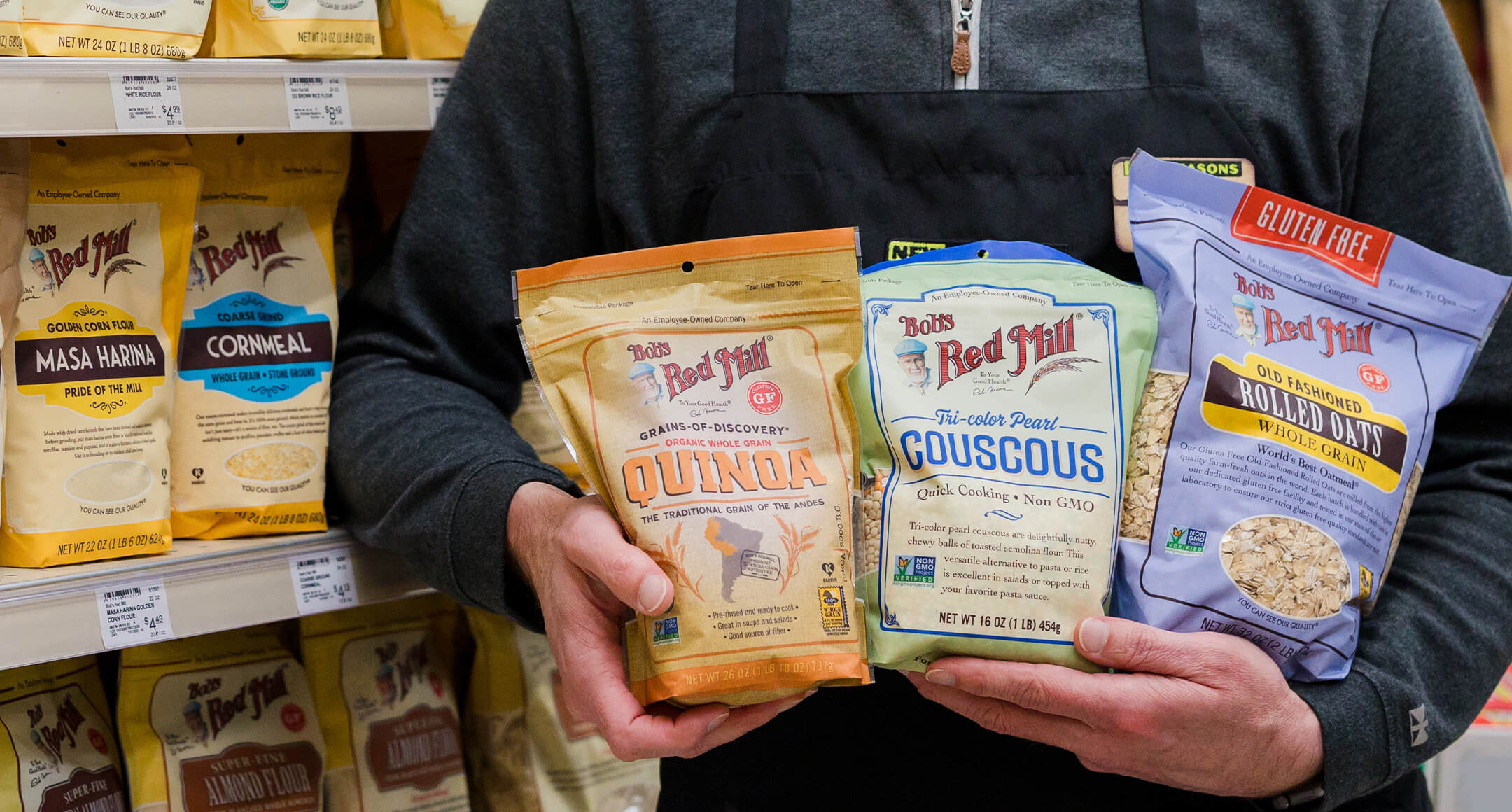 An individual holding bags of Bob’s Red Mill Quinoa, Rolled Oats, and Couscous. 
