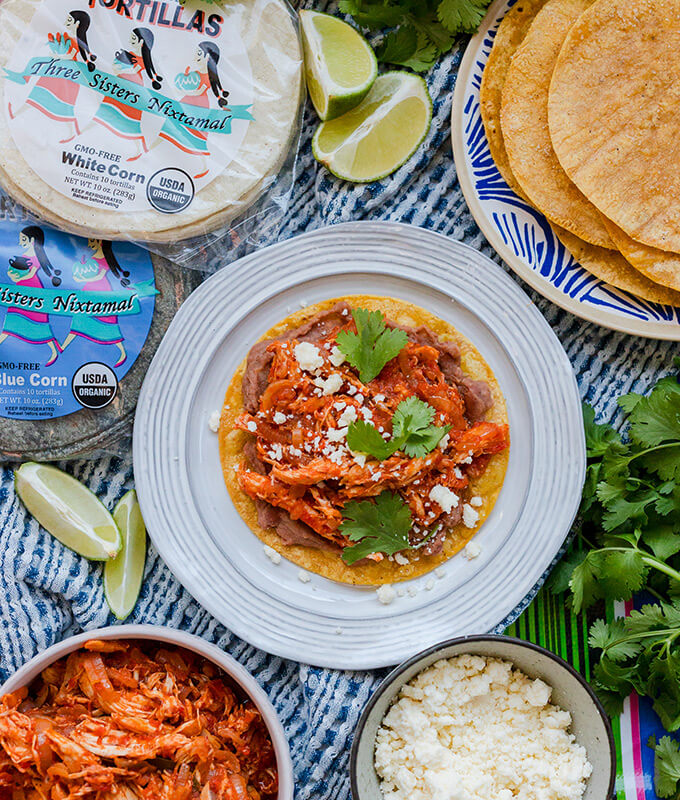 Three Sisters corn tortillas in a package and on a plate with chicken tinga forming a taco, topped with cojita cheese and cilantro.