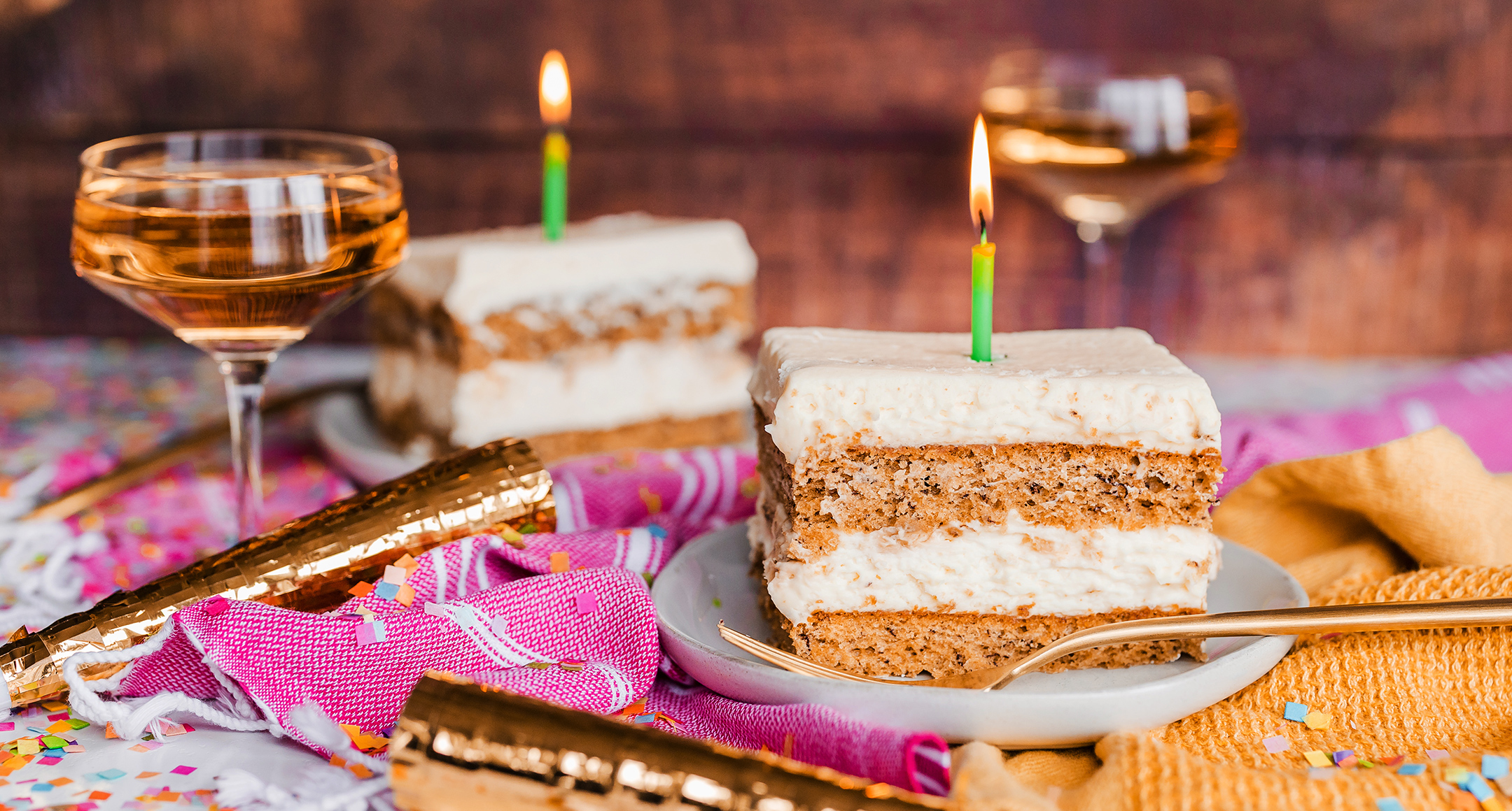 Two slices of birthday banana cake with lit candles sit atop a colorful tablecloth with a glass of wine. 