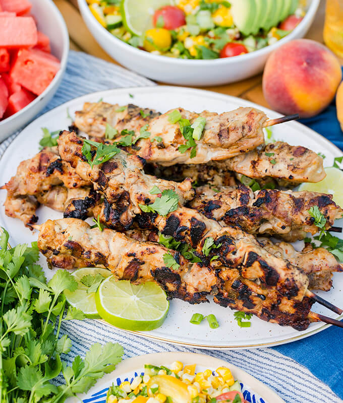 grilled meat skewers on a plate sprinkled with fresh herbs and with other picnic sides in the background