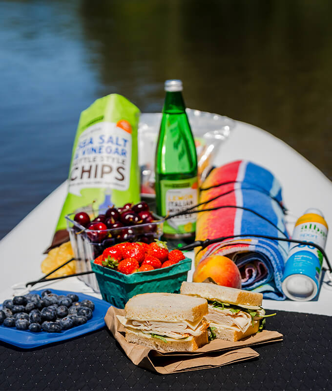 New Seasons branded items displayed at the end of a kayak along with fresh fruit, a sandwich and a towel.