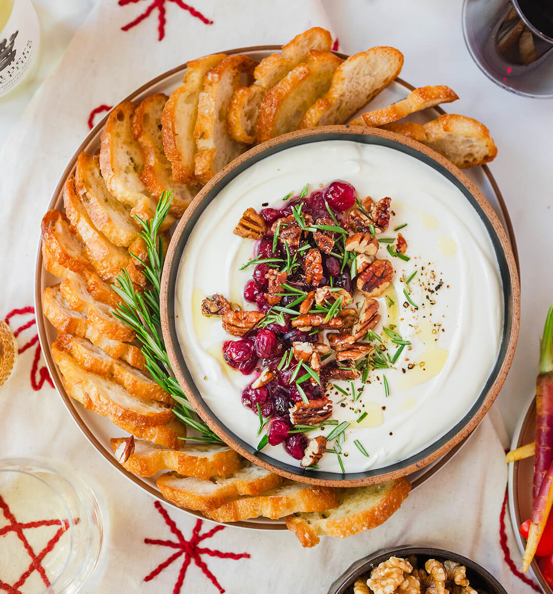 a bowl of whipped goat cheese topped with walnuts, cranberries and rosemary served with croustini